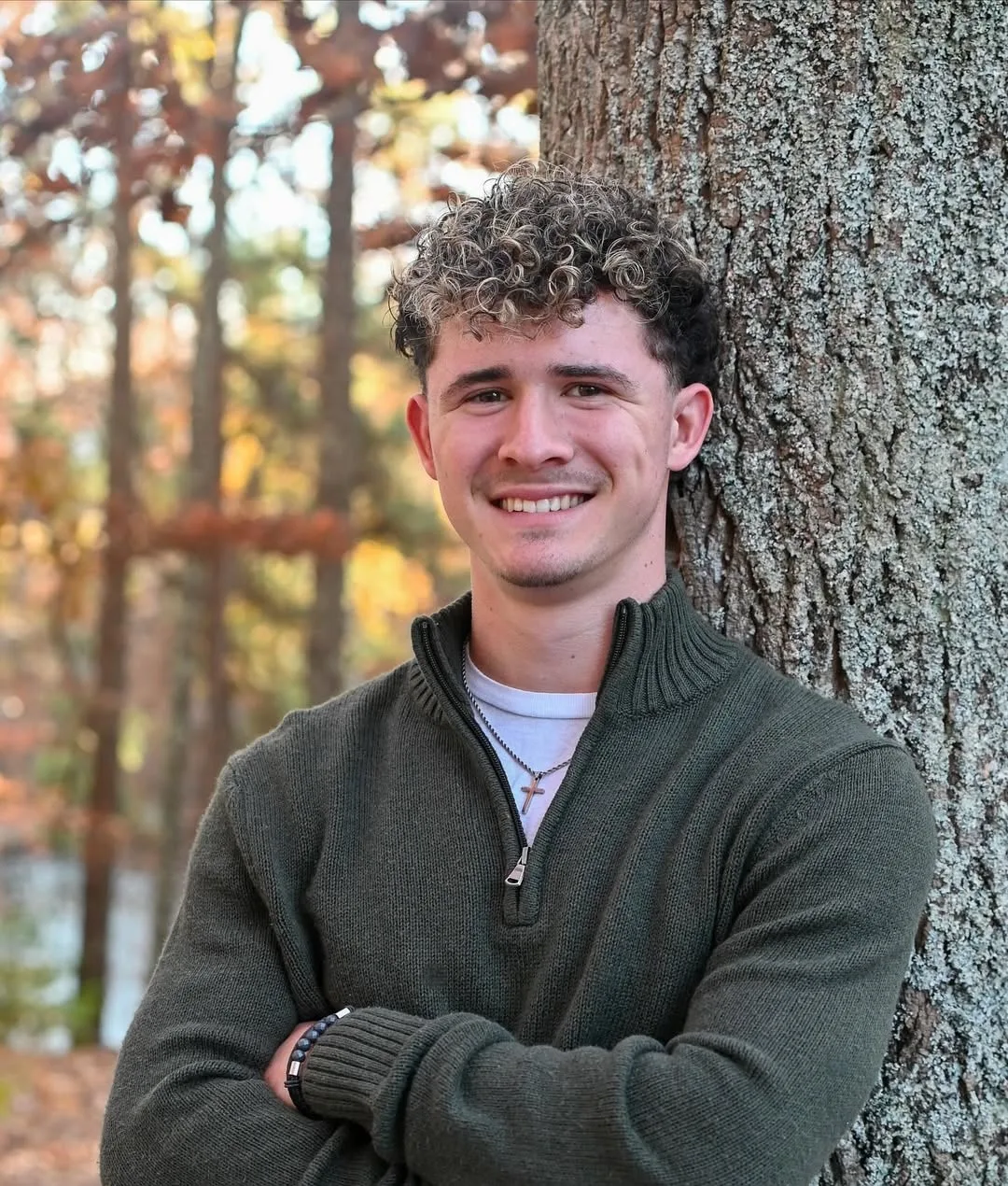 A young man in a green quarter-zip sweater leaning against a tree trunk in autumn