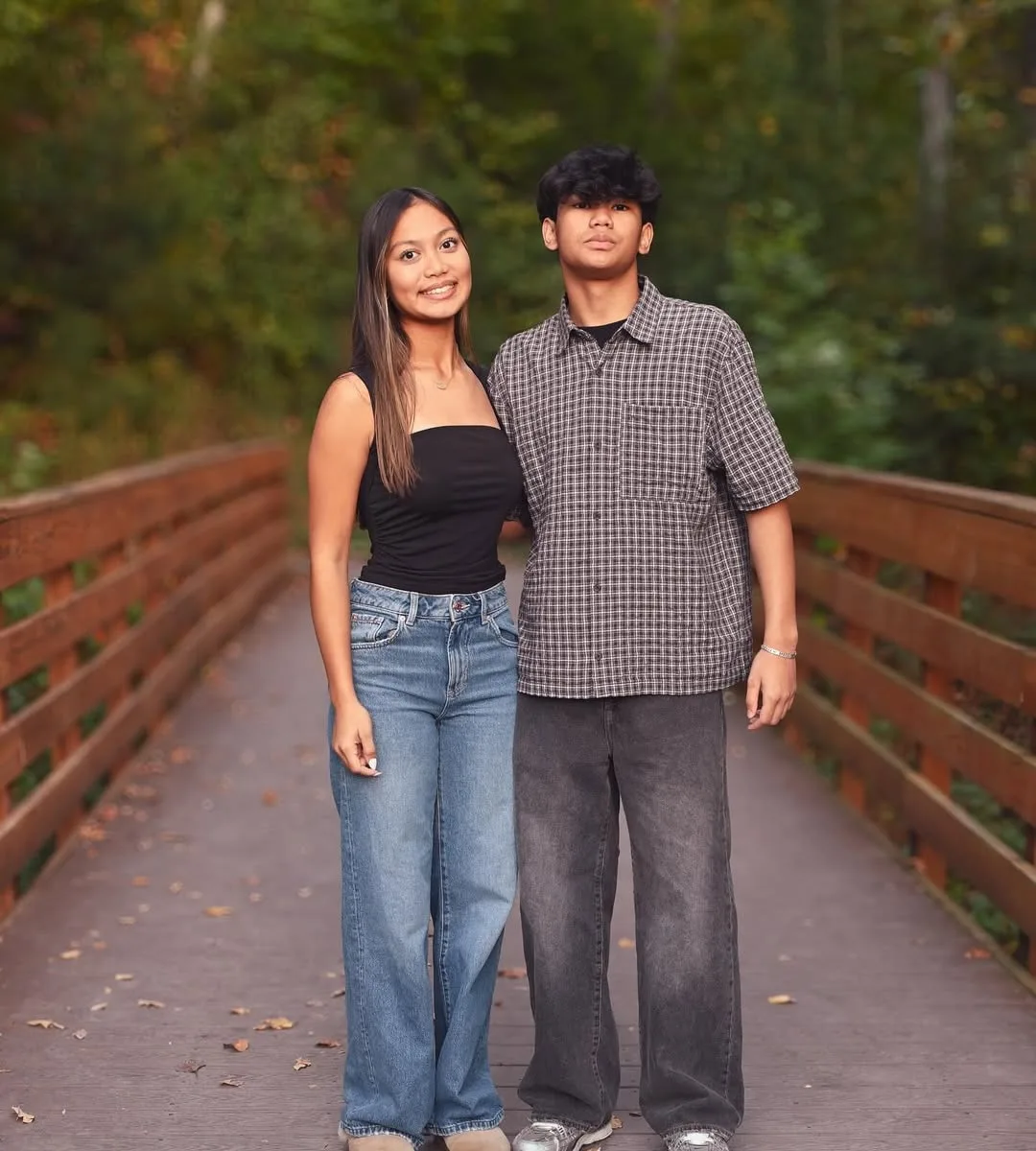 A brother and sister standing together on a wooden boardwalk lined with autumn trees